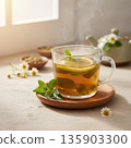 Hot herbal tea with lemon, mint leaves, and steam in a clear glass mug on a wooden coaster. 135903300