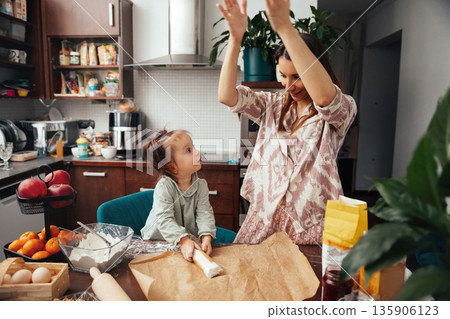 Mother and daughter bake together in a kitchen filled with ingredients and cooking tools while sharing moments of joy and creativity during the afternoon 135906123