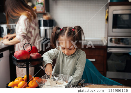 Child mixes ingredients while another person prepares food in a kitchen during a busy afternoon with fruit and cooking supplies around Child mixes ingredients while another person prepares food in a kitchen during a busy afternoon with fruit and cooking supplies around 135906126