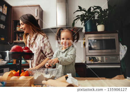 Mom and daughter bake together in a kitchen filled with fruits and cooking tools during daytime, creating sweet memories with smiles and laughter 135906127