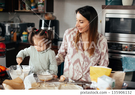 Woman and girl making dough in kitchen, enjoying time together, engaged in cooking activity while surrounded by baking ingredients Woman and girl making dough in kitchen, enjoying time together, engaged in cooking activity while surrounded by baking ingredients 135906132