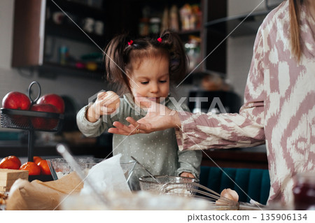 Child helps adult in kitchen with baking by handing eggs and preparing ingredients during a cooking session at home in the afternoon Child helps adult in kitchen with baking by handing eggs and preparing ingredients during a cooking session at home in the afternoon 135906134