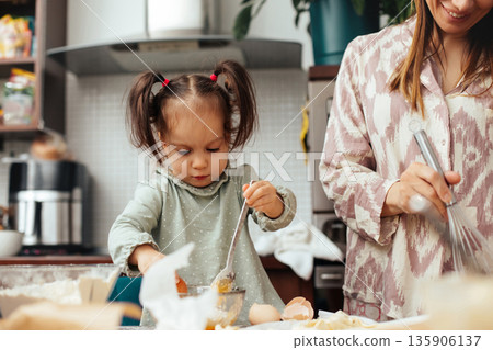 Child and adult cooking together in a kitchen while preparing food with various ingredients on the table around them during the daytime Child and adult cooking together in a kitchen while preparing food with various ingredients on the table around them during the daytime 135906137
