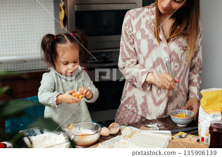 Mother and daughter bake together in a kitchen, making a cake and enjoying time together with ingredients and utensils spread out on the countertop 135906139