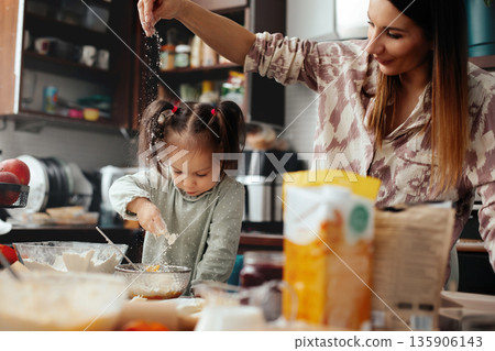 Mother and daughter cook together in the kitchen, making a dessert with flour and sugar on a busy countertop filled with cooking items during the afternoon Mother and daughter cook together in the kitchen, making a dessert with flour and sugar on a busy countertop filled with cooking items during the afternoon 135906143