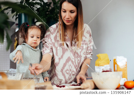 Mother and daughter prepare food together in a kitchen as they make pastries with various ingredients on the table Mother and daughter prepare food together in a kitchen as they make pastries with various ingredients on the table 135906147