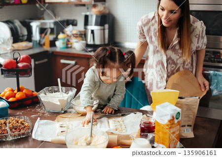 Child and adult working together in kitchen making baked goods with ingredients and utensils scattered around during daytime 135906161