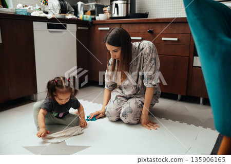 Mother cleans the floor while daughter plays in the kitchen during the day at home in a family setting 135906165