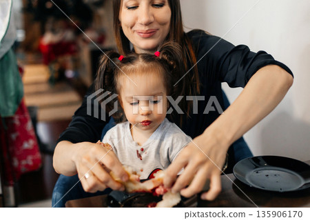 Mother helps daughter eat food while sitting at a table in a home setting during daytime with casual clothes and smiles 135906170