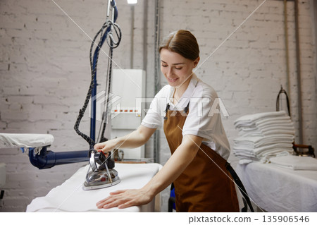 A woman worker wearing an apron is skillfully ironing a shirt on a table 135906546