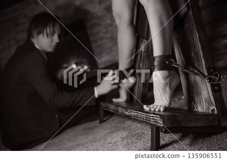 A man chains a woman's legs to a BDSM cross. A man chains a woman's legs to a BDSM cross. 135906551