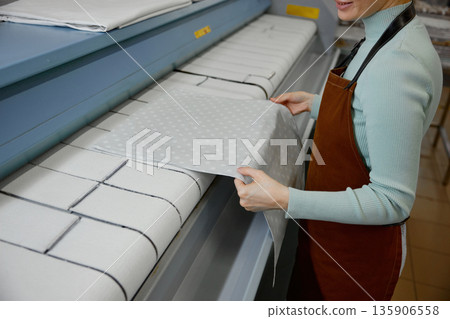 A young woman worker is ironing towels on a table in a laundromat 135906558