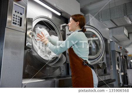 A woman worker is loading various clothes into a washing machine 135906568