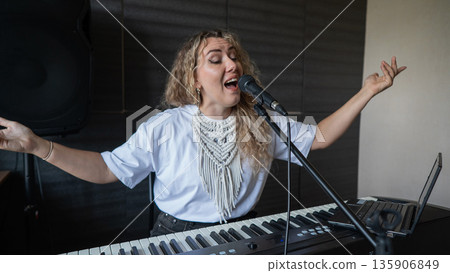 Caucasian woman singing into microphone and playing synthesizer and recording herself on laptop. Caucasian woman singing into microphone and playing synthesizer and recording herself on laptop. 135906849