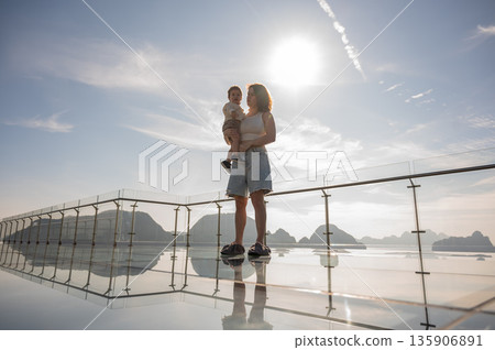 Caucasian woman holds her little son in her arms while standing on a glass bridge in the mountains. Caucasian woman holds her little son in her arms while standing on a glass bridge in the mountains. 135906891