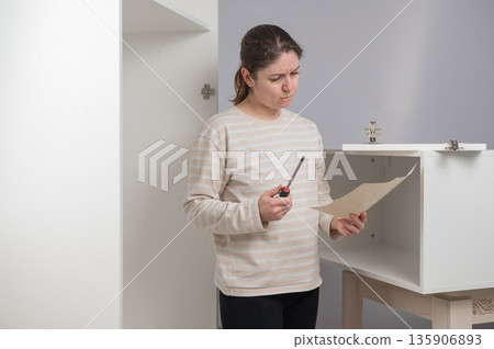 A Caucasian woman looks at the assembly instructions for a wardrobe with confusion. A Caucasian woman looks at the assembly instructions for a wardrobe with confusion. 135906893