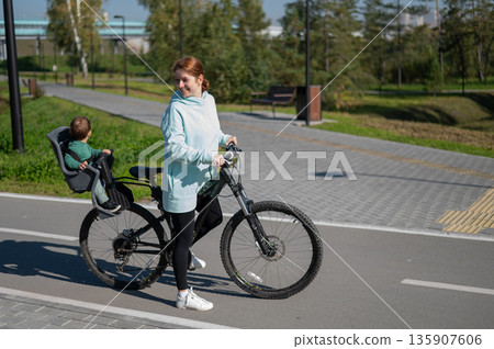 Caucasian woman rides bicycle with one-year-old son in park. Caucasian woman rides bicycle with one-year-old son in park. 135907606