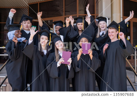 Happy students of different nationalities in graduation gowns hold their diplomas. 135907688