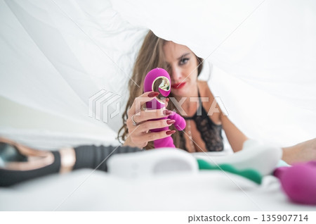 Woman in lace lingerie holds a dildo. A beautiful Caucasian woman lies under the sheets and holds a dildo. Woman in lace lingerie holds a dildo. A beautiful Caucasian woman lies under the sheets and holds a dildo. 135907714