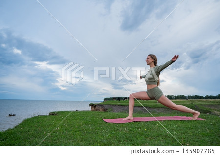 Caucasian woman doing yoga on the river bank. Warrior pose. Caucasian woman doing yoga on the river bank. Warrior pose. 135907785