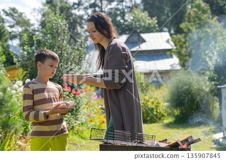 Caucasian woman grilling sausages on a barbecue with her son.  135907845