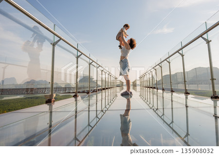 Caucasian woman holds her little son in her arms while standing on a glass bridge in the mountains.  135908032
