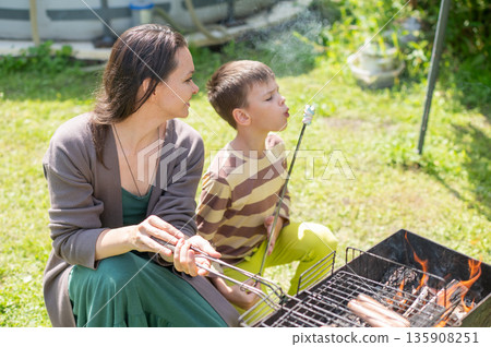 Caucasian woman grilling sausages on a barbecue with her son. Caucasian woman grilling sausages on a barbecue with her son. 135908251