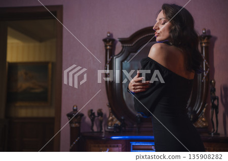A beautiful brunette in a black evening dress poses in front of a retro-style mirror. A beautiful brunette in a black evening dress poses in front of a retro-style mirror. 135908282