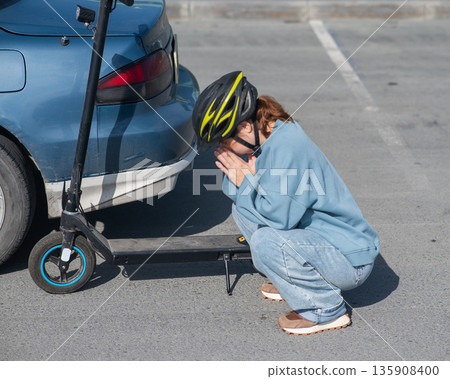 A distressed woman examines the damage after a collision between an electric scooter and a car. A distressed woman examines the damage after a collision between an electric scooter and a car. 135908400