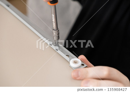 Person's hand uses screwdriver to install a white furniture drawer runner, symbolizing home assembly, practical repair, and a focused DIY project 135908427