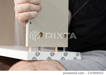 Persons hands joining new flat-pack furniture panels together with dowels and cam locks, creating custom storage for a modern home interior 135908435