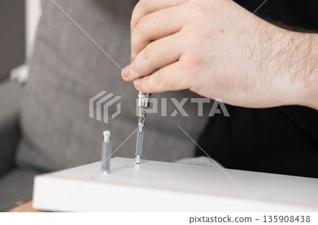 Male hand utilizing a screwdriver to assemble white flat-pack furniture, symbolizing home improvement, practical repair, and do-it-yourself project completion Male hand utilizing a screwdriver to assemble white flat-pack furniture, symbolizing home improvement, practical repair, and do-it-yourself project completion 135908438