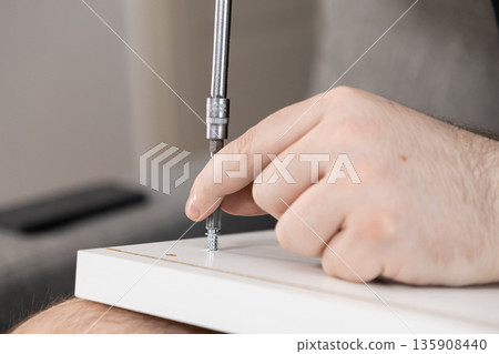 Person's hand using a screwdriver to fasten a metal bolt into a white furniture panel, representing practical home assembly and personal DIY project work Person's hand using a screwdriver to fasten a metal bolt into a white furniture panel, representing practical home assembly and personal DIY project work 135908440