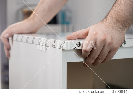 Man's hands carefully positioning a white flat-pack furniture panel, featuring protective PE-LD recycling symbols on its edge, symbolizing a home assembly or DIY improvement project Man's hands carefully positioning a white flat-pack furniture panel, featuring protective PE-LD recycling symbols on its edge, symbolizing a home assembly or DIY improvement project 135908443