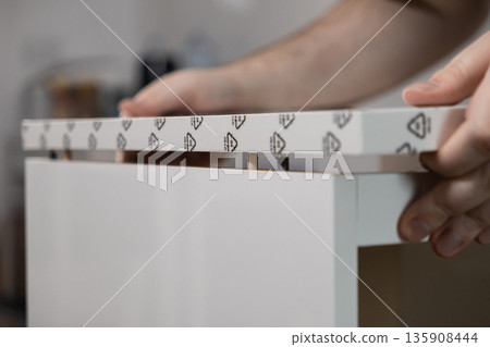 Close-up of person's hands carefully assembling white modular flat-pack furniture components using wooden dowels and pins, emphasizing a DIY home improvement project Close-up of person's hands carefully assembling white modular flat-pack furniture components using wooden dowels and pins, emphasizing a DIY home improvement project 135908444