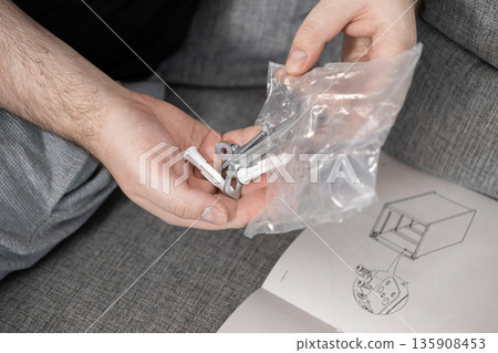 Person's hands organize diverse furniture assembly hardware from plastic bag, positioned near instruction manual, ready for home project installation Person's hands organize diverse furniture assembly hardware from plastic bag, positioned near instruction manual, ready for home project installation 135908453