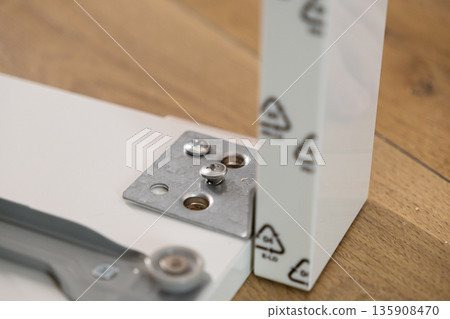 White furniture components featuring a metal bracket and partially fastened screw, illustrating a hands-on do-it-yourself assembly project for home renovation White furniture components featuring a metal bracket and partially fastened screw, illustrating a hands-on do-it-yourself assembly project for home renovation 135908470
