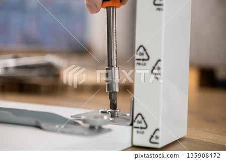 Person's hand using a screwdriver to assemble a white furniture panel with a metal bracket, highlighting practical home improvement and do-it-yourself tasks 135908472