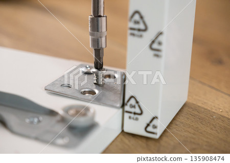 Screwdriver bit precisely fastening a shiny metal screw into a sturdy bracket on white furniture, symbolizing hands-on home improvement and successful furniture assembly 135908474