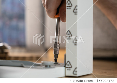 A human hand expertly uses a screwdriver to fasten a small screw into a white furniture panel, embodying efficient home assembly and practical DIY projects A human hand expertly uses a screwdriver to fasten a small screw into a white furniture panel, embodying efficient home assembly and practical DIY projects 135908478