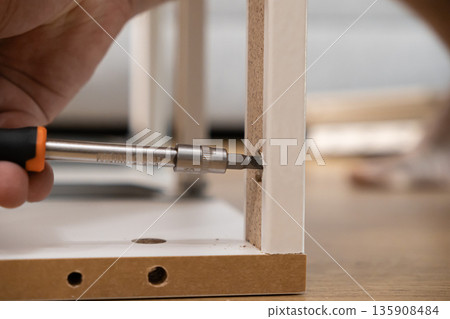 Person's hand actively using a screwdriver to meticulously assemble new white flat-pack furniture, embodying a focus on practical home improvement and do-it-yourself tasks 135908484