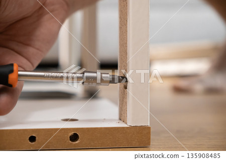 Person's hand skillfully using a screwdriver to assemble white particle board furniture, representing practical DIY home improvement and craftsmanship Person's hand skillfully using a screwdriver to assemble white particle board furniture, representing practical DIY home improvement and craftsmanship 135908485