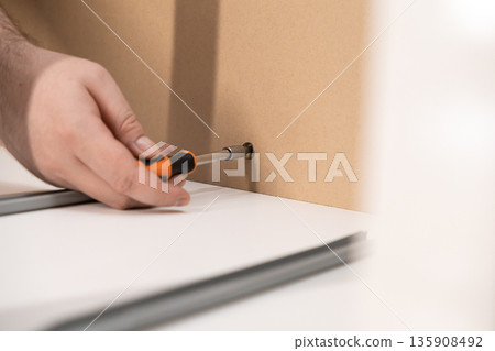 Person's hand expertly using an orange screwdriver to securely fasten a screw into a light brown wooden furniture panel, symbolizing DIY assembly 135908492