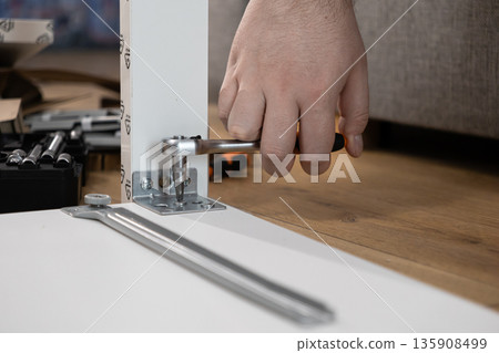 Close-up of hand assembling modern white modular furniture with a ratchet screwdriver, fastening a metal bracket, representing practical home improvement and DIY work Close-up of hand assembling modern white modular furniture with a ratchet screwdriver, fastening a metal bracket, representing practical home improvement and DIY work 135908499