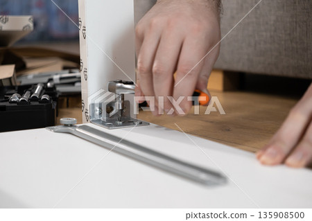 A person's hands skillfully using a ratchet screwdriver to securely fasten a metal bracket onto a white furniture panel, illustrating home assembly and practical DIY projects A person's hands skillfully using a ratchet screwdriver to securely fasten a metal bracket onto a white furniture panel, illustrating home assembly and practical DIY projects 135908500