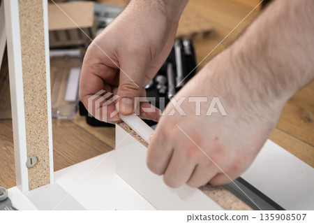 Person's hands carefully inserting white plastic dowel into modern particle board furniture, symbolizing home assembly, DIY skill, and interior design setup Person's hands carefully inserting white plastic dowel into modern particle board furniture, symbolizing home assembly, DIY skill, and interior design setup 135908507