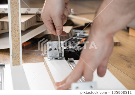 Close-up of hands diligently assembling furniture, fastening a metal bracket with a screwdriver onto wooden panel, representing practical DIY home improvement skill Close-up of hands diligently assembling furniture, fastening a metal bracket with a screwdriver onto wooden panel, representing practical DIY home improvement skill 135908511