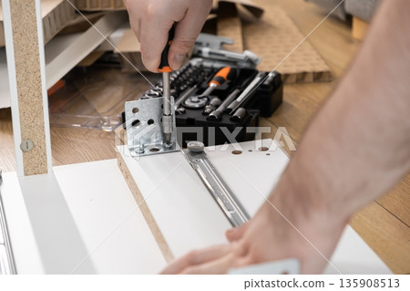 Close-up of a person's hands actively using a screwdriver to securely attach metal hardware during flat-pack furniture assembly, symbolizing home improvement and DIY effort Close-up of a person's hands actively using a screwdriver to securely attach metal hardware during flat-pack furniture assembly, symbolizing home improvement and DIY effort 135908513