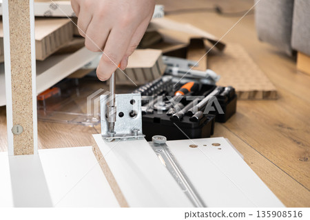 Person's hand fastening a metal bracket with a screwdriver during flat-pack furniture assembly, representing practical home improvement and DIY renovation projects Person's hand fastening a metal bracket with a screwdriver during flat-pack furniture assembly, representing practical home improvement and DIY renovation projects 135908516
