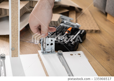 A person's hand meticulously using a screwdriver to assemble white flat-pack furniture, symbolizing DIY home improvement and repair projects A person's hand meticulously using a screwdriver to assemble white flat-pack furniture, symbolizing DIY home improvement and repair projects 135908517